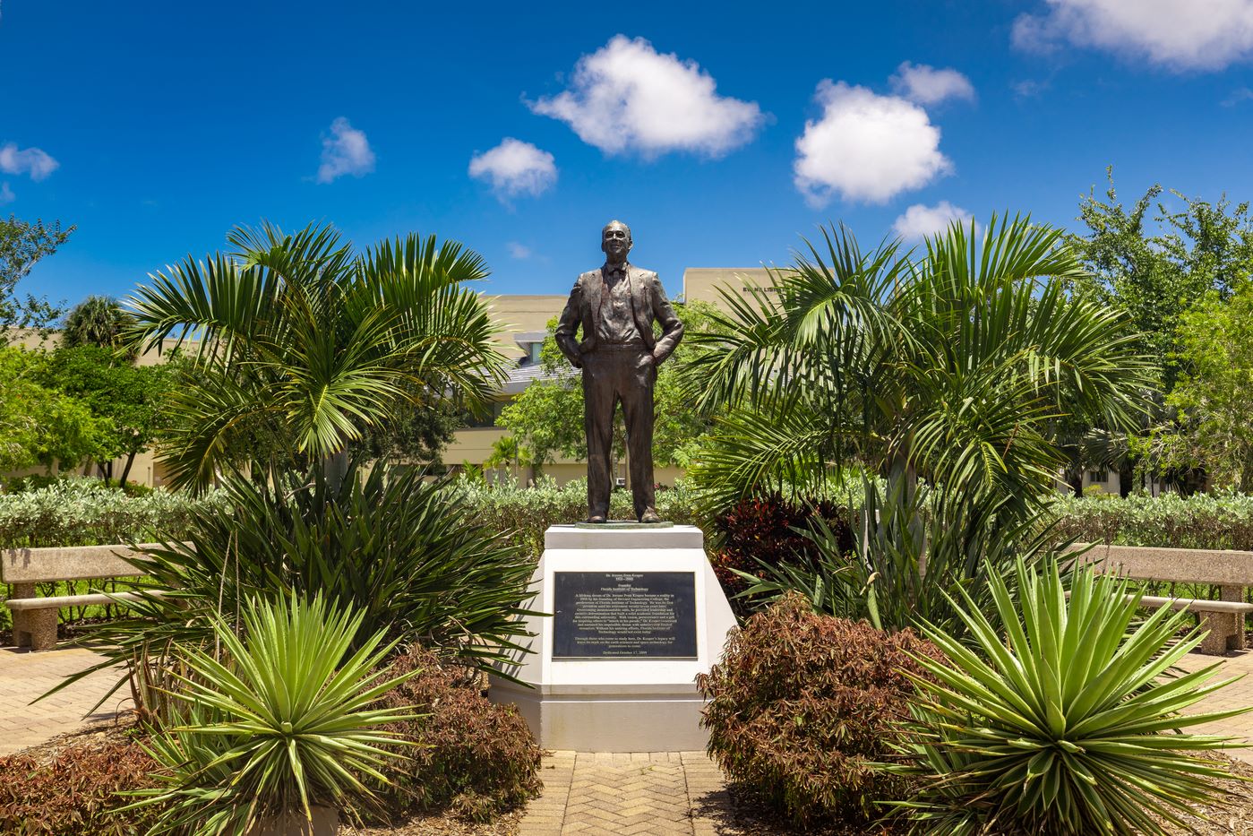 Statue of a distinguished man in a suit, surrounded by a lush tropical garden with palm trees under a clear blue sky.